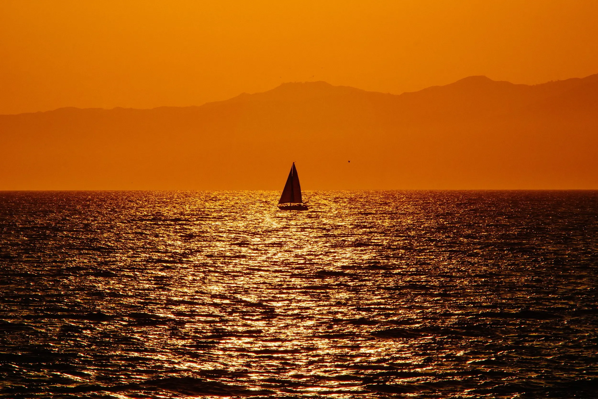 A sailboat near the horizon on the black ocean water, silhouetted against mountains. Golden hour.