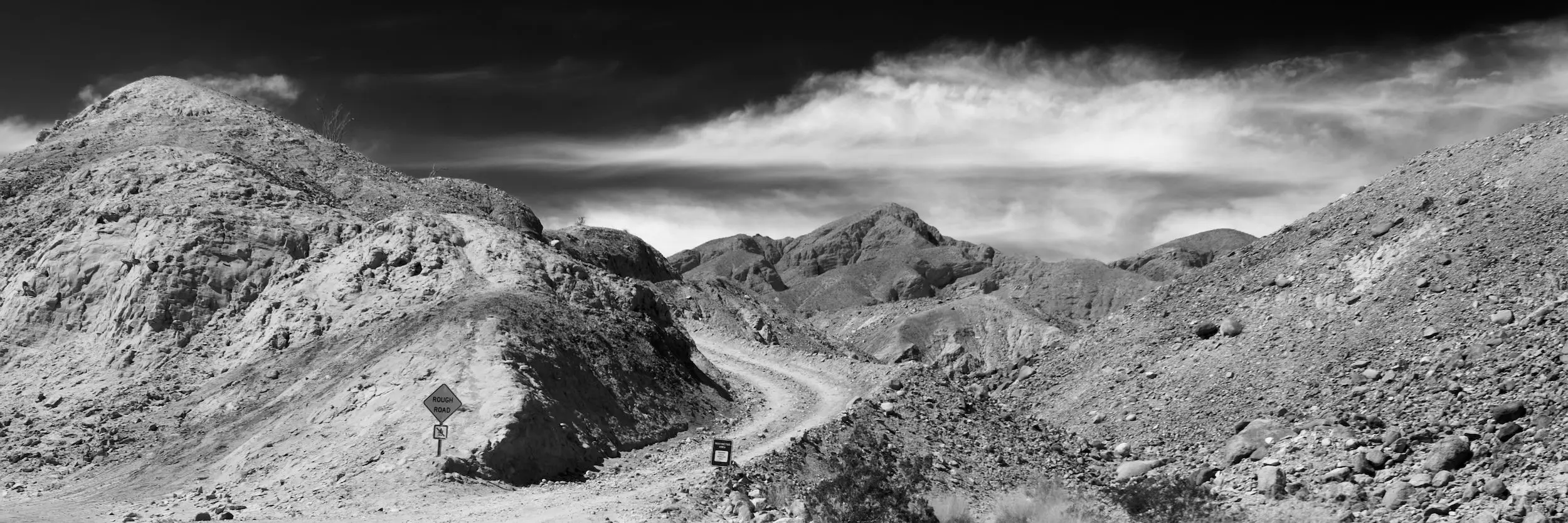 Black and white wide shot of a curving road in between a rocky landscape with dramatic clouds in the sky.