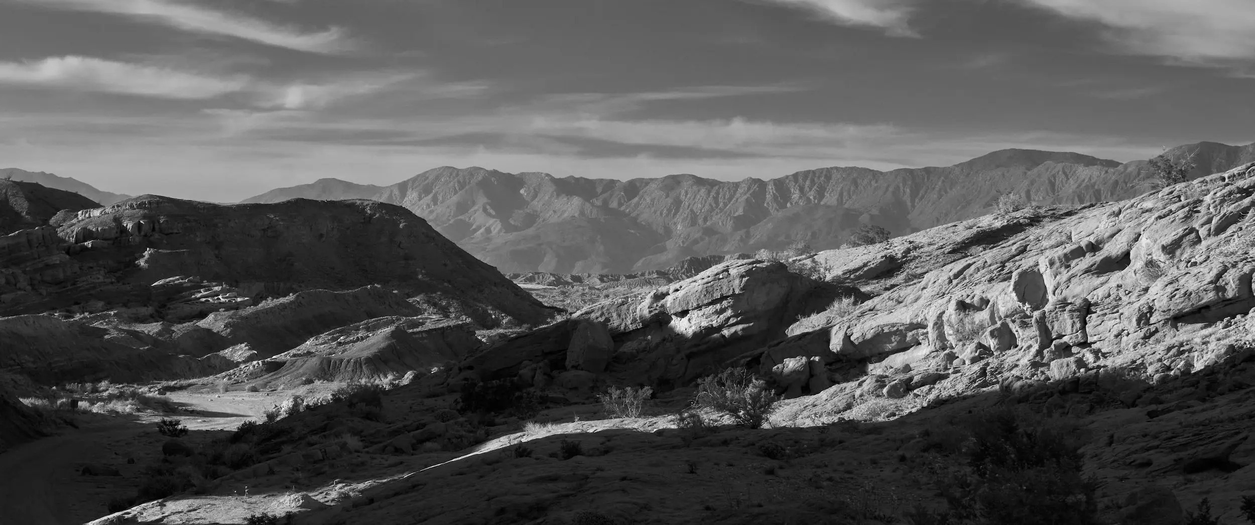 Black and white wide shot with sunset light and shadow raking through a rocky landscape with mountains in the background.